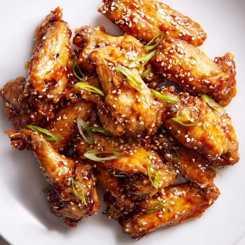 Golden brown Asian Sticky Wings coated in a sticky, glossy soy-ginger glaze on a serving platter with sesame seeds and sliced green onions.