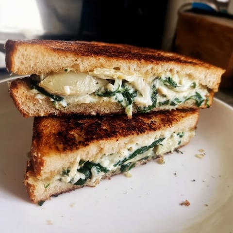 Two vegetarian Spinach Artichoke Grilled Cheese halves on a plate, the crisp sourdough revealing a vibrant green and cheesy interior.