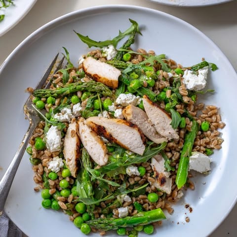 A close-up of Warm Spring Farro Chicken Salad, showcasing juicy sliced chicken, tender farro grains, bright green asparagus, and peas tossed in a zesty lemon vinaigrette.